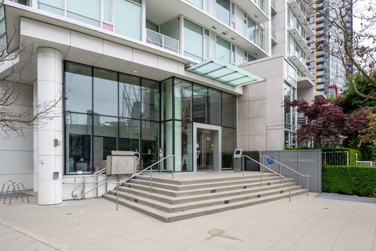 Modern building entrance with glass doors, metal railings, and steps leading up to 1205 West Georgia, Vancouver. A glass canopy and building sign are visible near landscaped greenery.