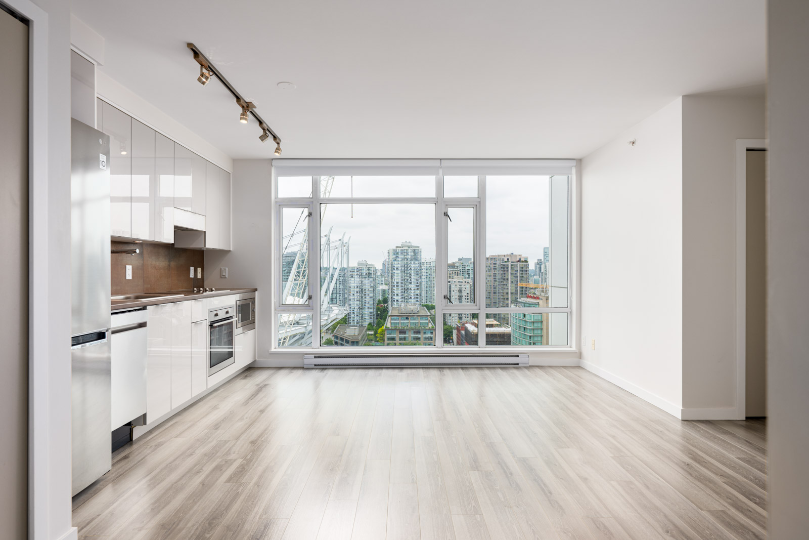 Modern, unfurnished apartment with white kitchen, stainless steel appliances, light wood floors, and large windows offering a city view.