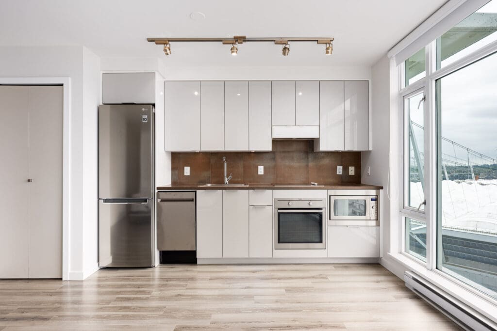 Modern kitchen with white cabinets, stainless steel appliances, wood flooring, large window, and track lighting.