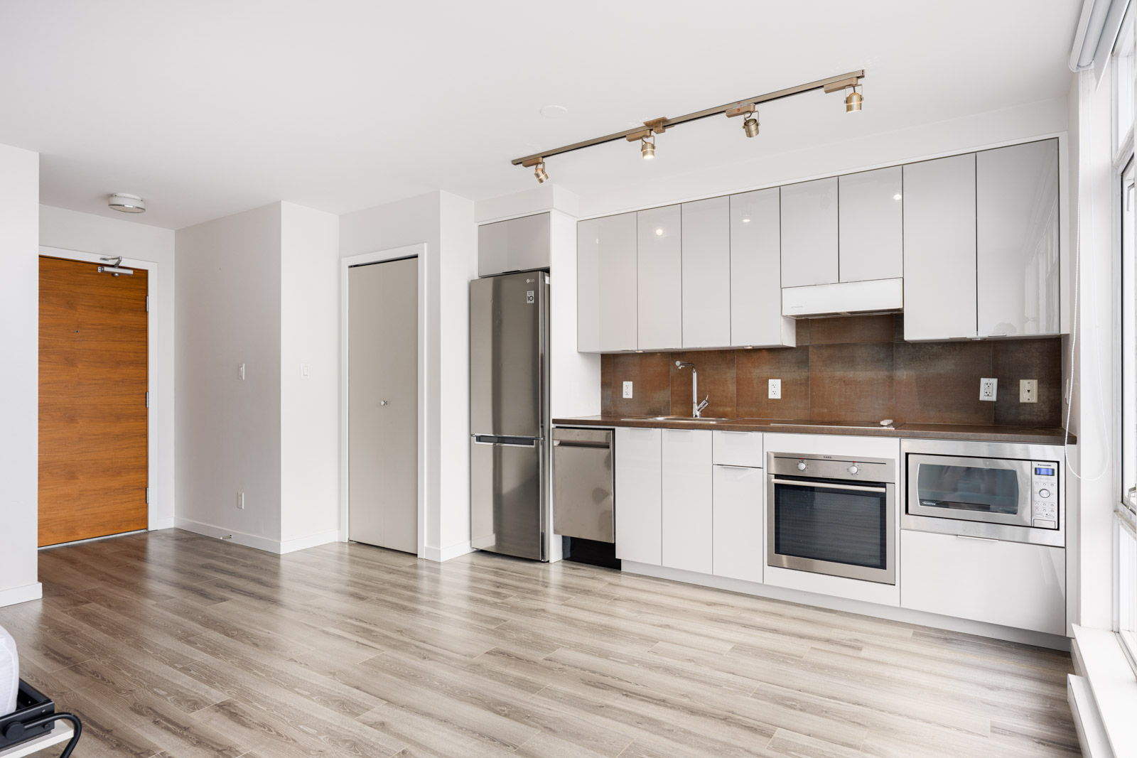 Modern apartment kitchen with white cabinets, stainless steel appliances, wood flooring, and track lighting, adjacent to a wooden entrance door and a closet.