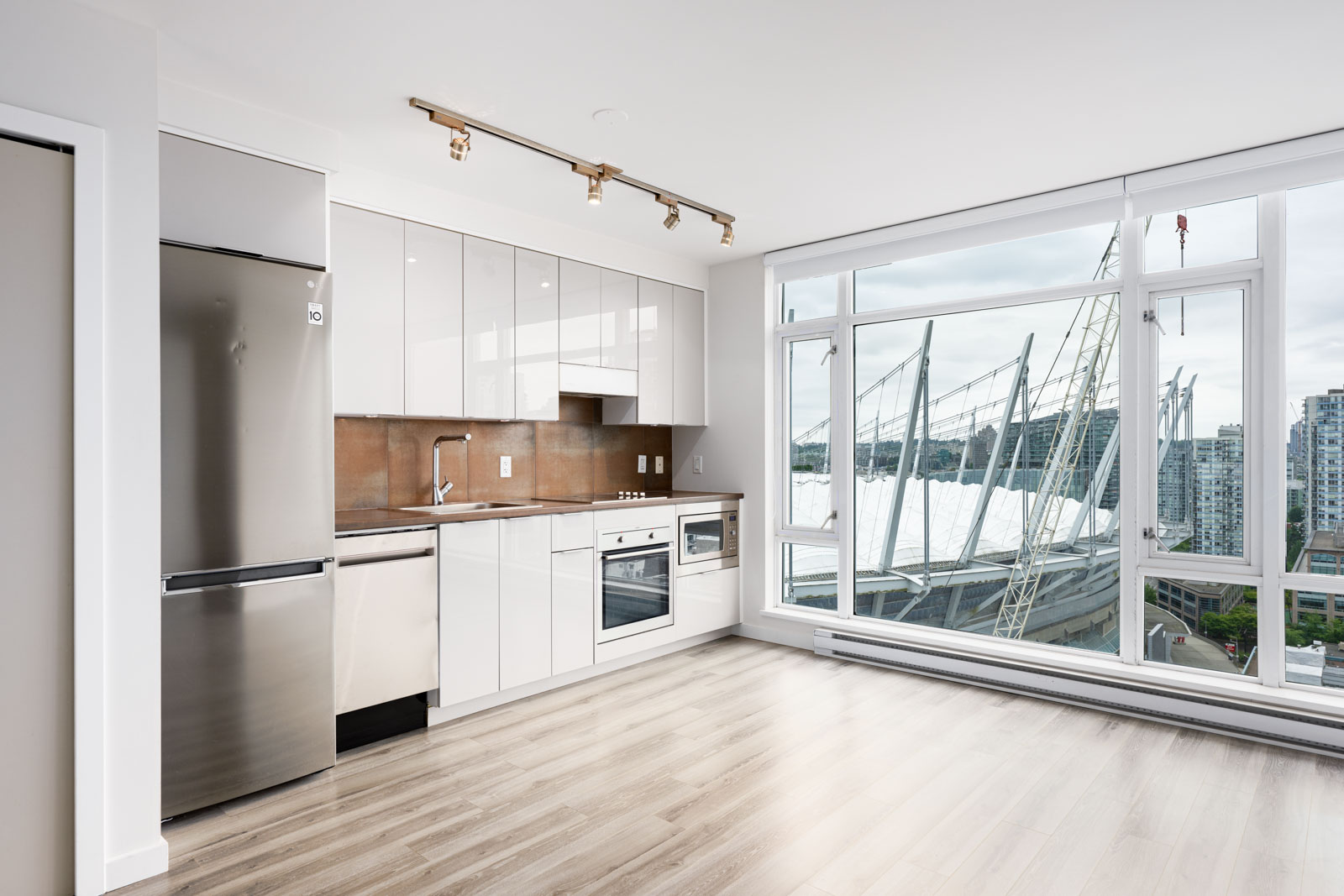 Modern apartment kitchen with stainless steel appliances, white cabinets, wood floors, and large windows offering a city view with stadium and buildings.