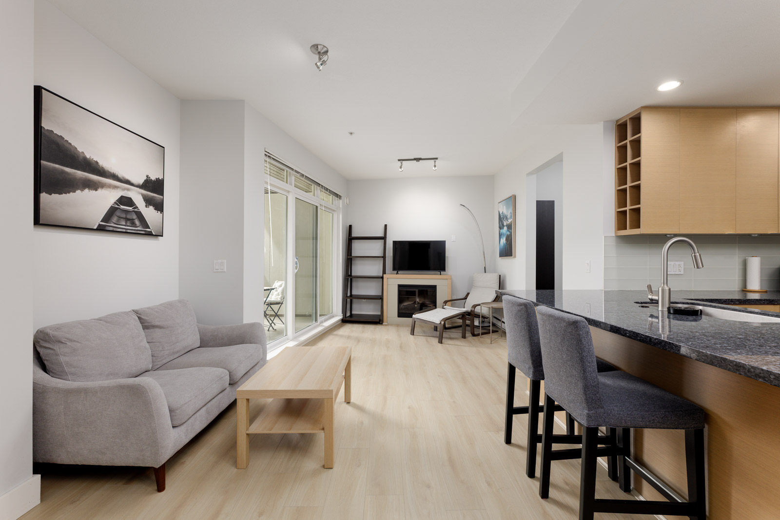 Modern living room and kitchen with light wood flooring, gray sofa, wooden coffee table, bar stools, TV, and large window letting in natural light.