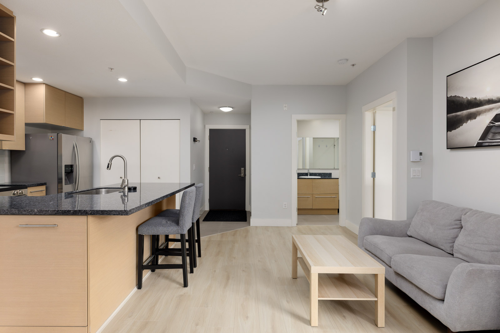 Modern apartment interior with kitchen, island seating, gray sofa, light wood coffee table, and visible bathroom vanity in the background.