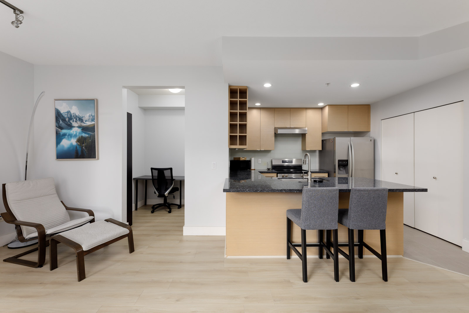 Modern kitchen with light wood cabinets, black countertops, and stainless steel appliances, adjacent to a seating area with two chairs and a small workspace in the background.