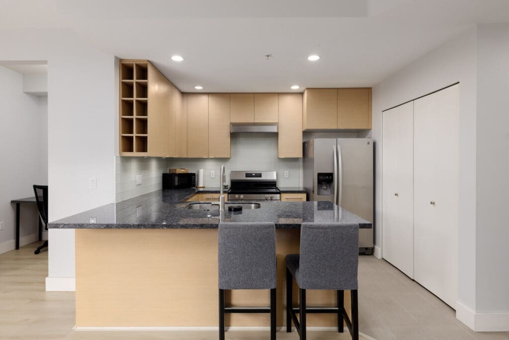 Modern kitchen with light wood cabinets, black granite countertop, stainless steel appliances, and two gray upholstered bar stools at the island.