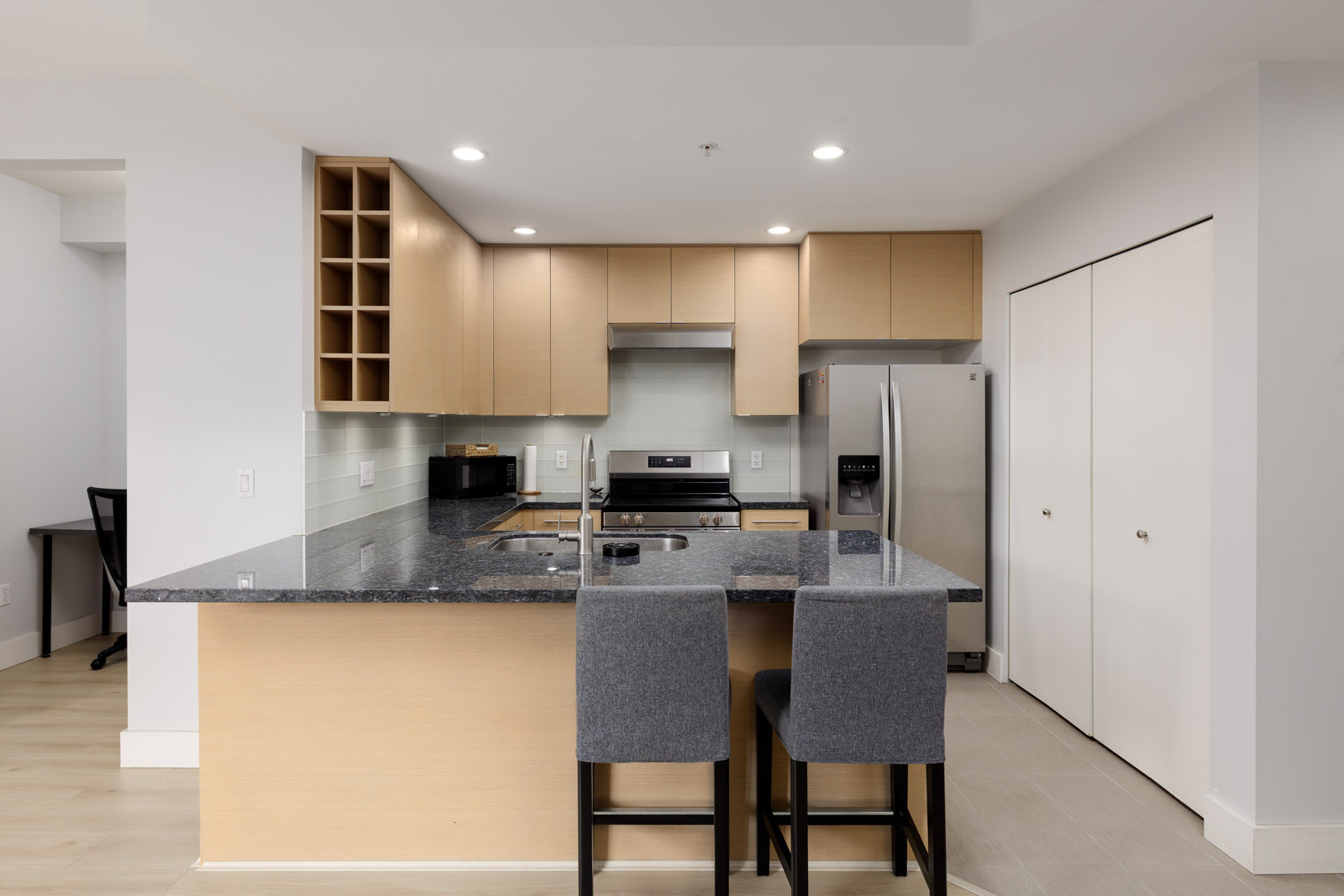 Modern kitchen with light wood cabinets, black granite countertop, stainless steel appliances, and two gray upholstered bar stools at the island.