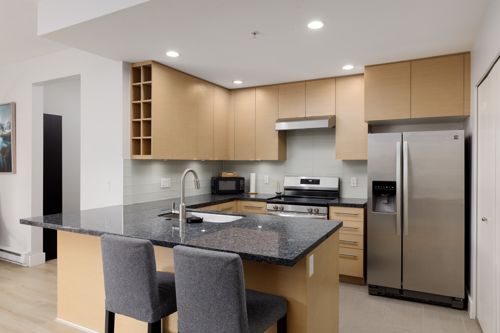 Modern kitchen with light wood cabinets, stainless steel appliances, a black granite island countertop, two gray chairs, and under-cabinet lighting.