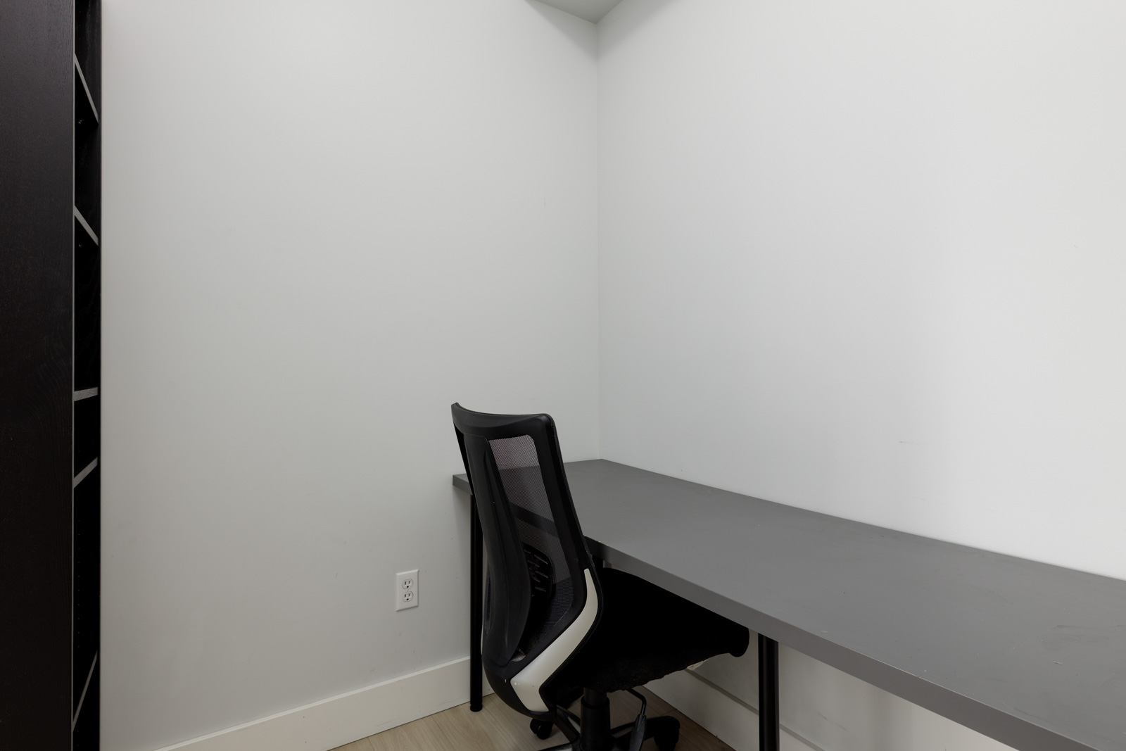 A small, minimalist office space with a black office chair at a gray desk against white walls and a power outlet visible on the left wall.