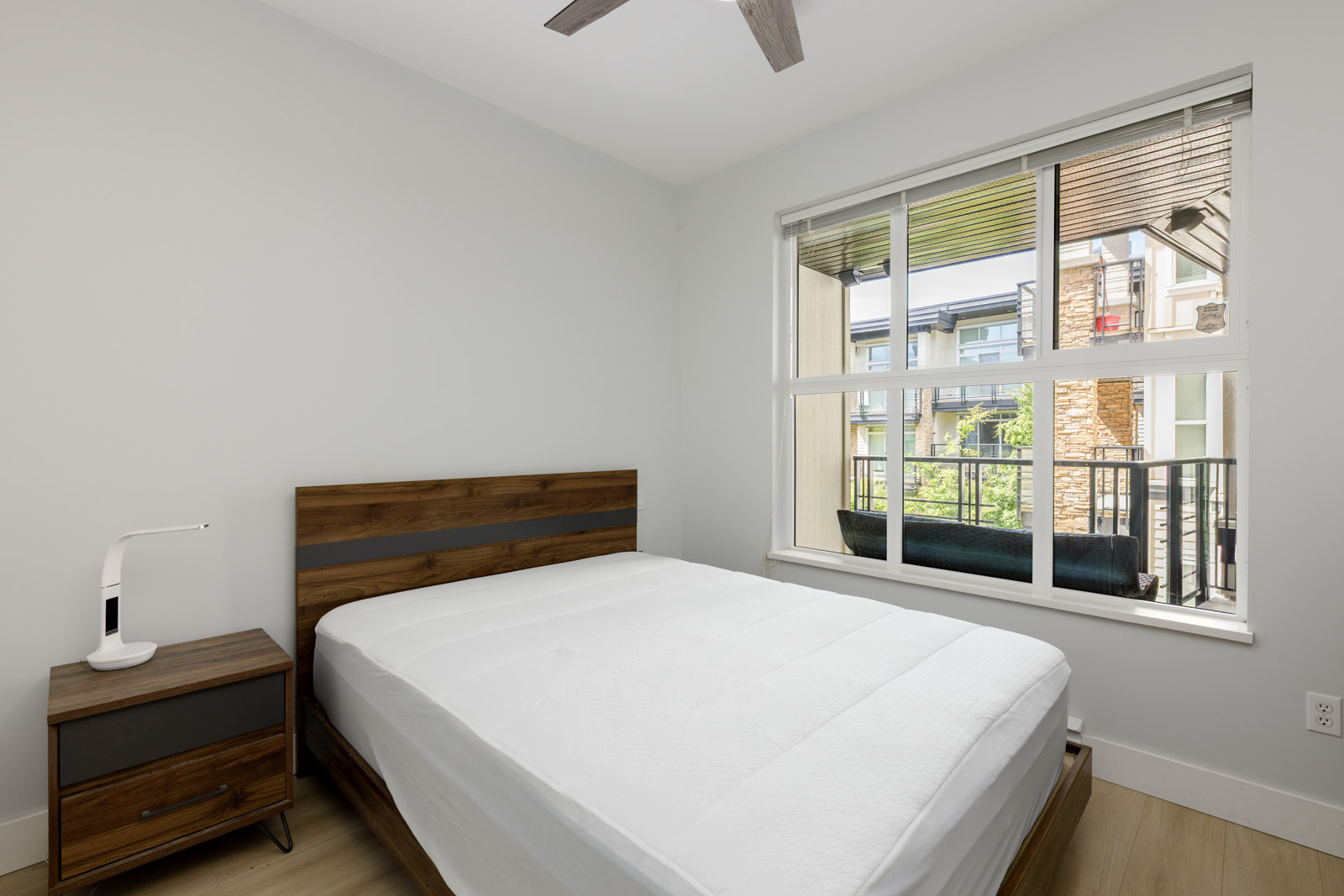 Minimalist bedroom with a wooden bed frame, white bedding, a nightstand with a small lamp, large window, and light-colored walls and flooring.