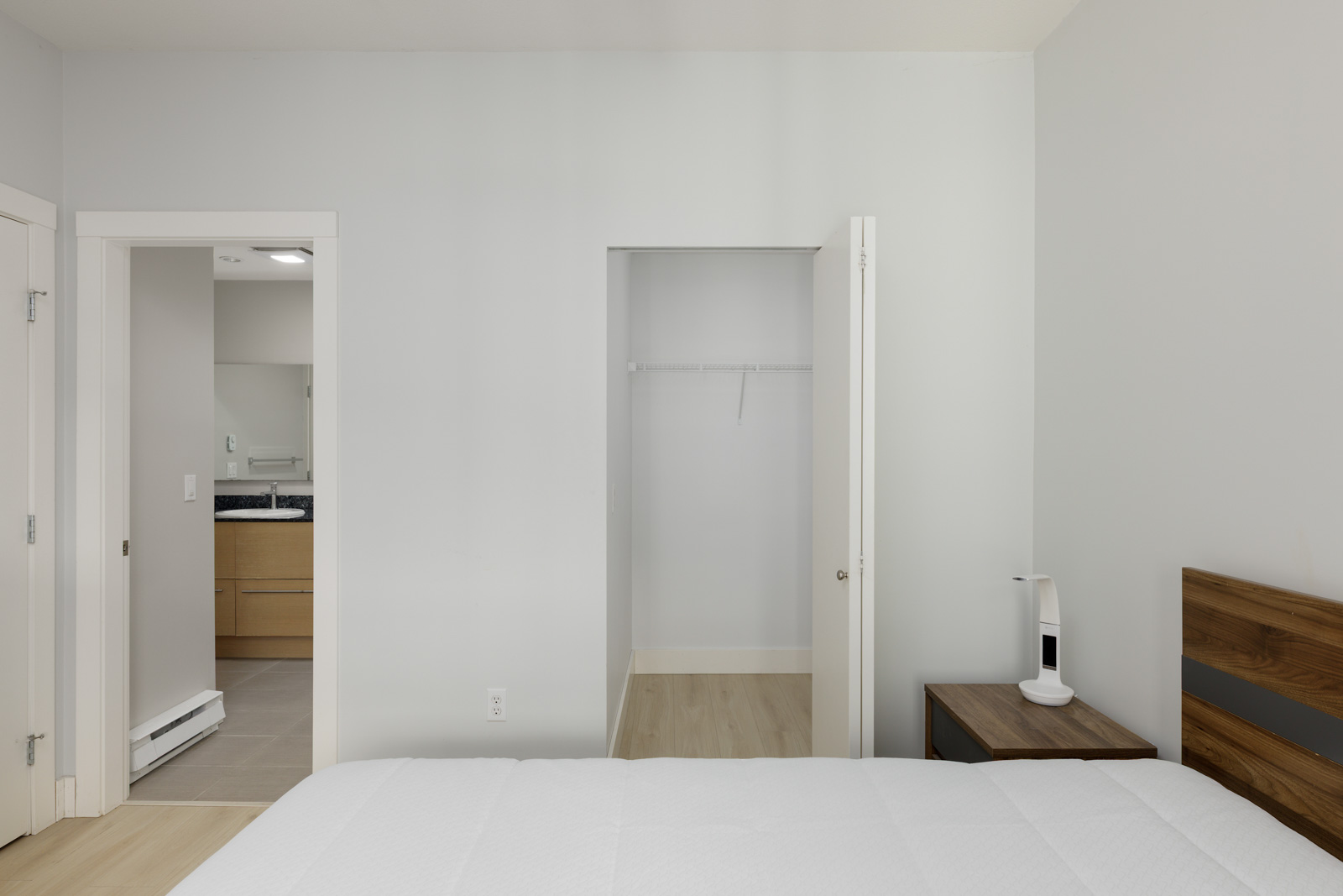 A minimalist bedroom with a bed in the foreground, an empty open closet, a nightstand, and a view into a bathroom with a wooden vanity.