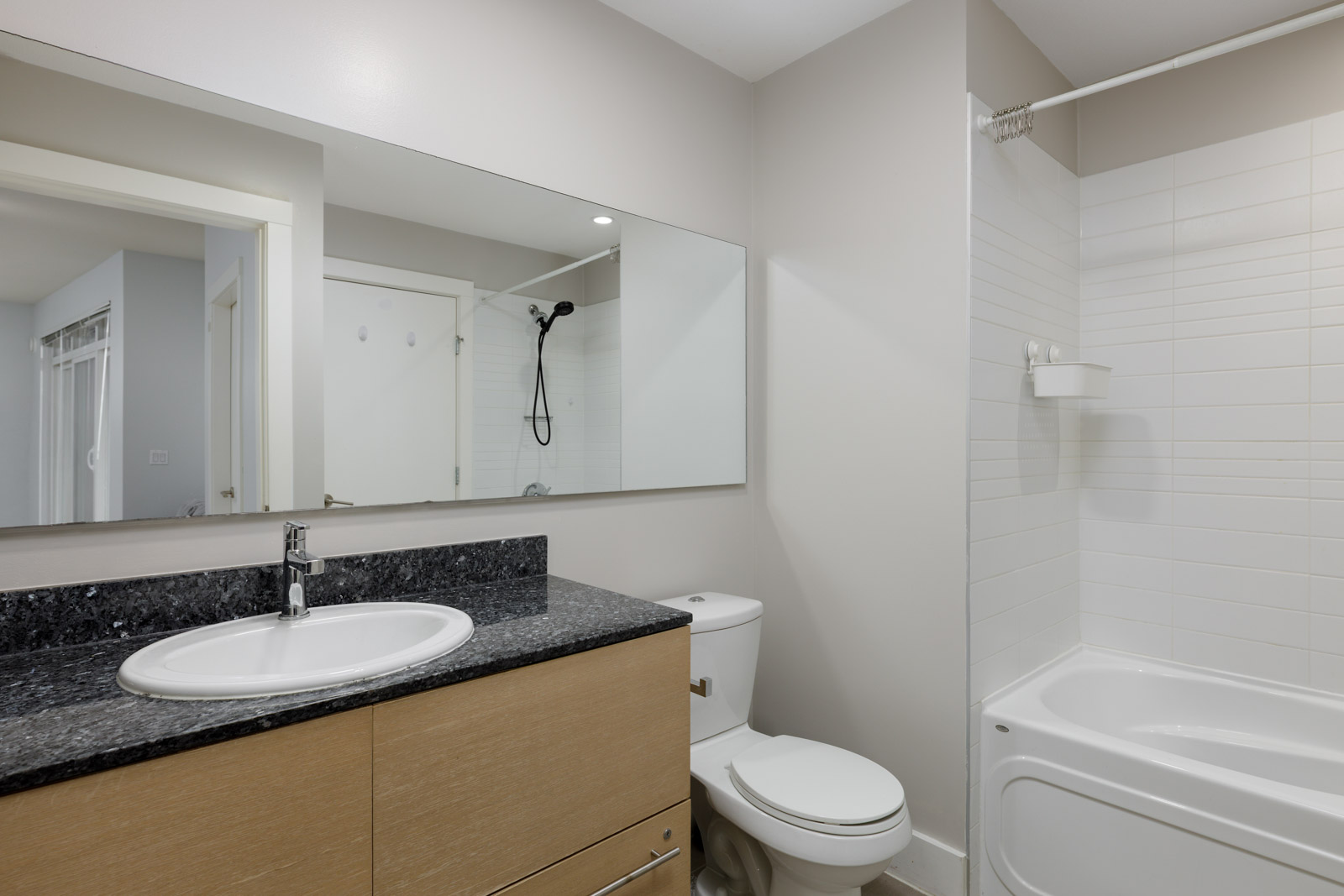 Modern bathroom with a wood vanity, granite countertop, sink, toilet, large wall mirror, and a white bathtub with a showerhead.