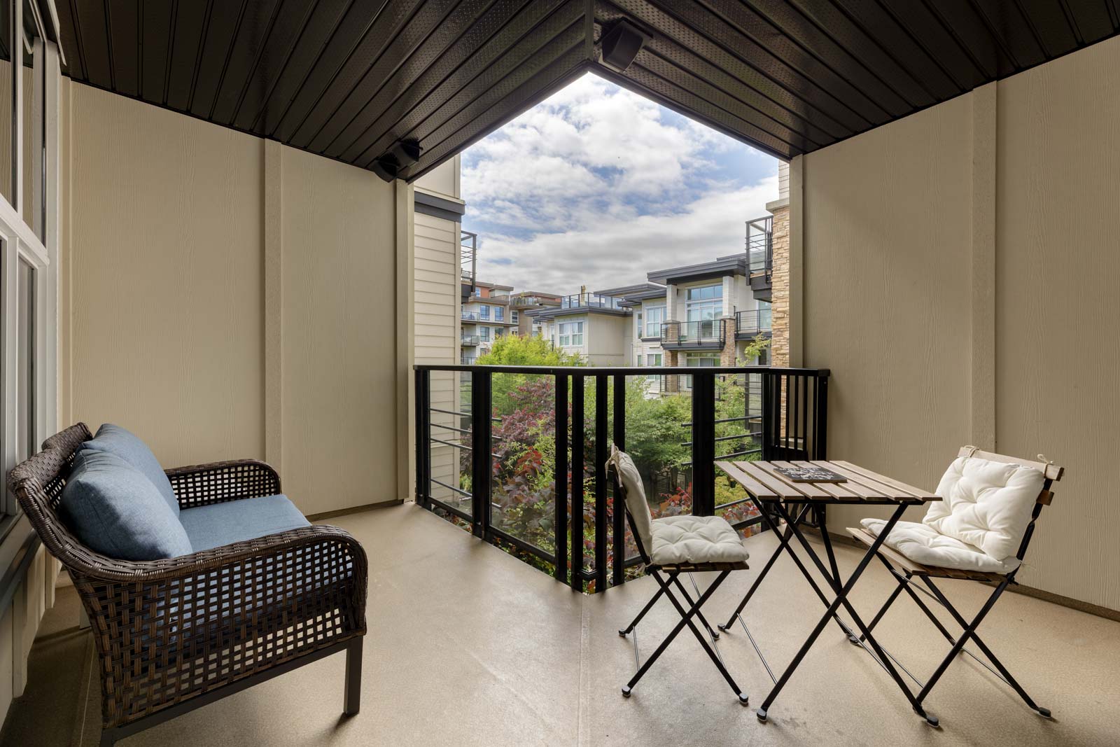Covered balcony with a wicker loveseat, a small table, two chairs, and a view of neighboring buildings and greenery under a partly cloudy sky.