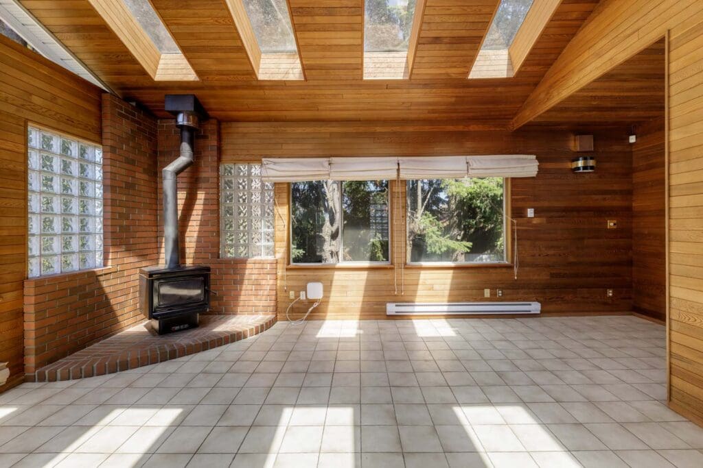 A sunlit room with wood-paneled walls, large windows, skylights, a tile floor, and a wood stove with a metal chimney on a brick hearth.