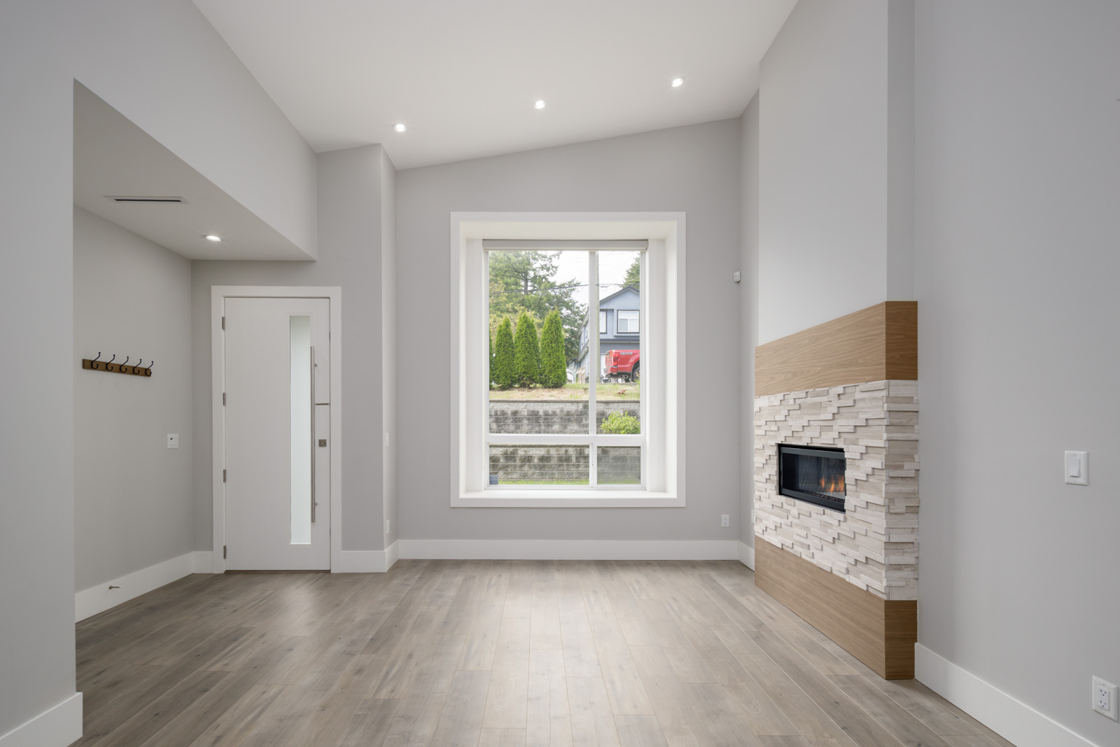 Modern empty living room with light wood flooring, large window, built-in fireplace with stone surround, white walls, and recessed ceiling lights.