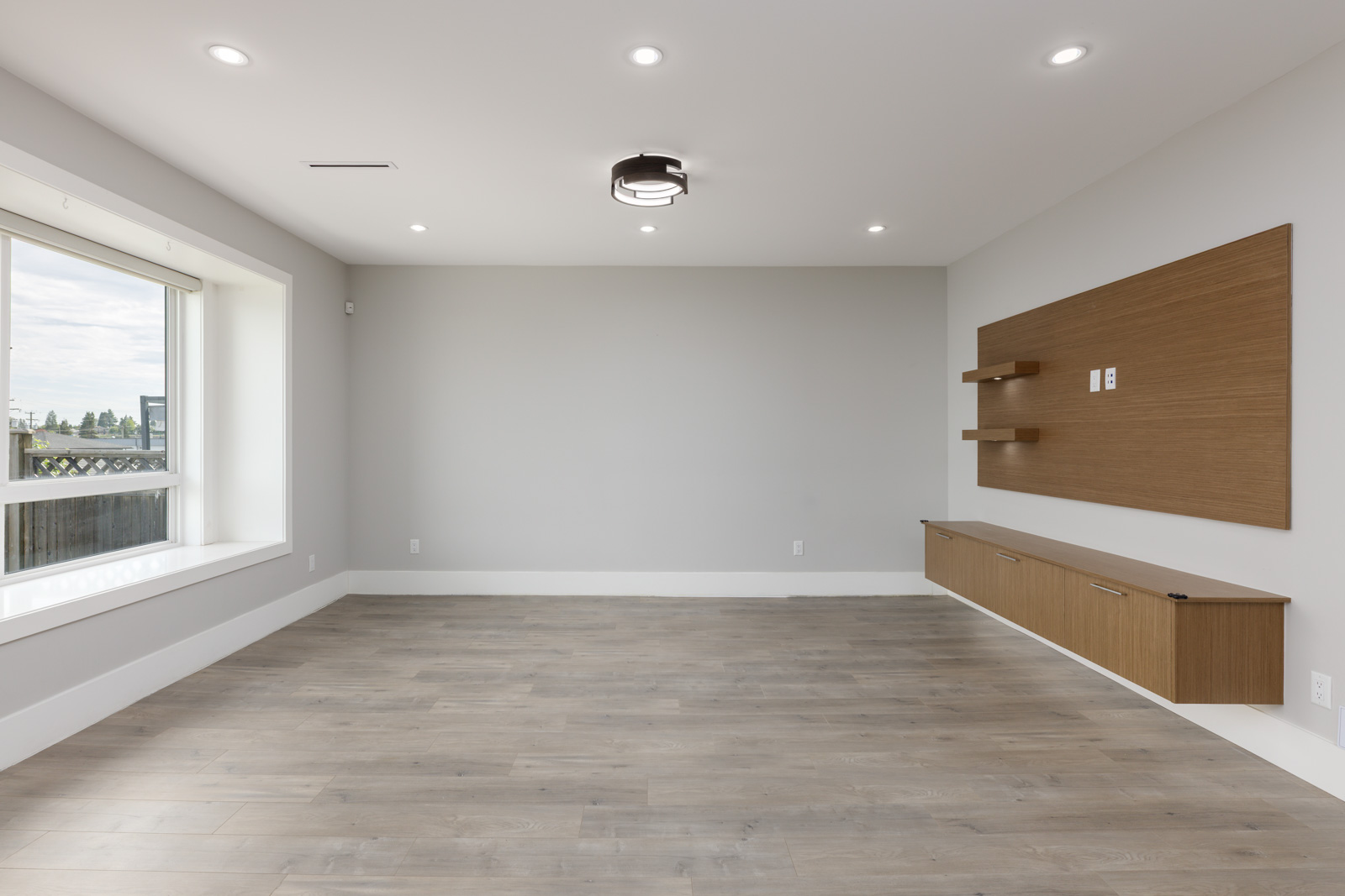 Empty modern room with light wood flooring, large window, recessed lighting, and a wall-mounted wooden storage unit with shelves.