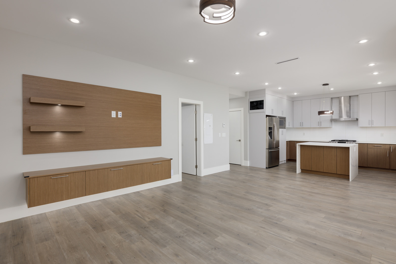 Modern, unfurnished kitchen and living area with wood floors, white cabinets, built-in oven, and wall-mounted shelves and cabinets. Recessed ceiling lights illuminate the space.