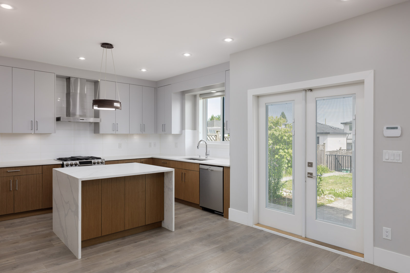 Modern kitchen with white and wood cabinets, center island, stainless steel appliances, large window over sink, and double glass doors leading to a backyard.