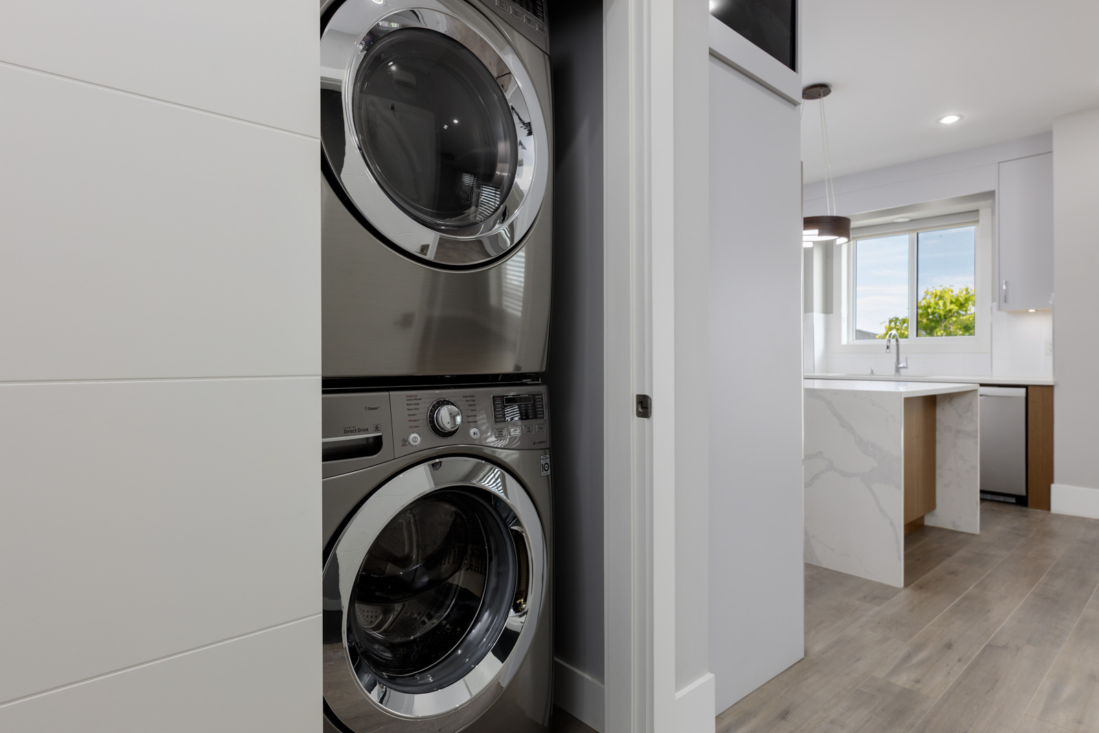 Stacked washer and dryer units in a closet next to a modern kitchen with light wood floors and white cabinetry.