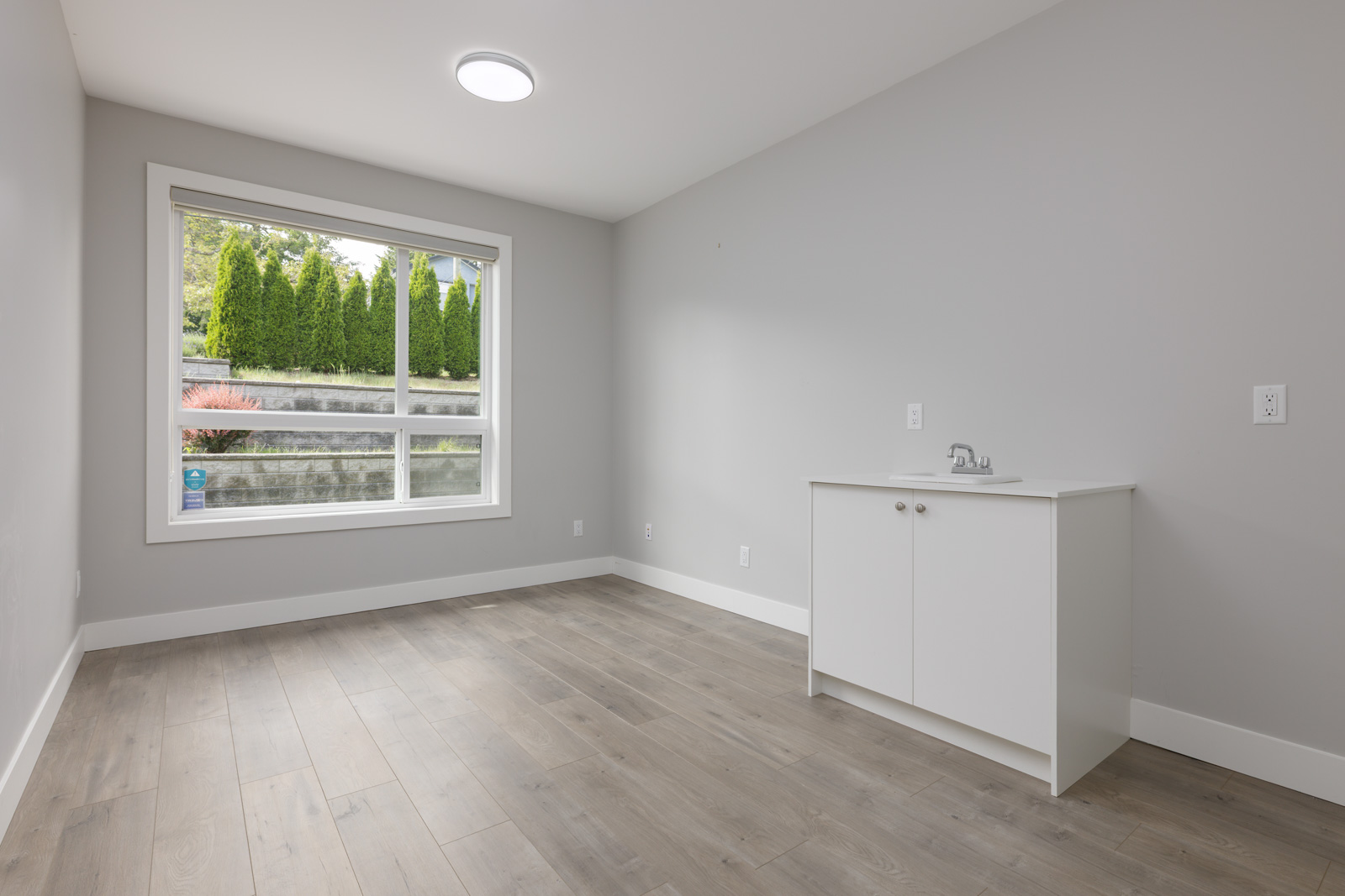 Empty room with light gray walls, wood flooring, a large window, and a small white cabinet with a sink in the corner.