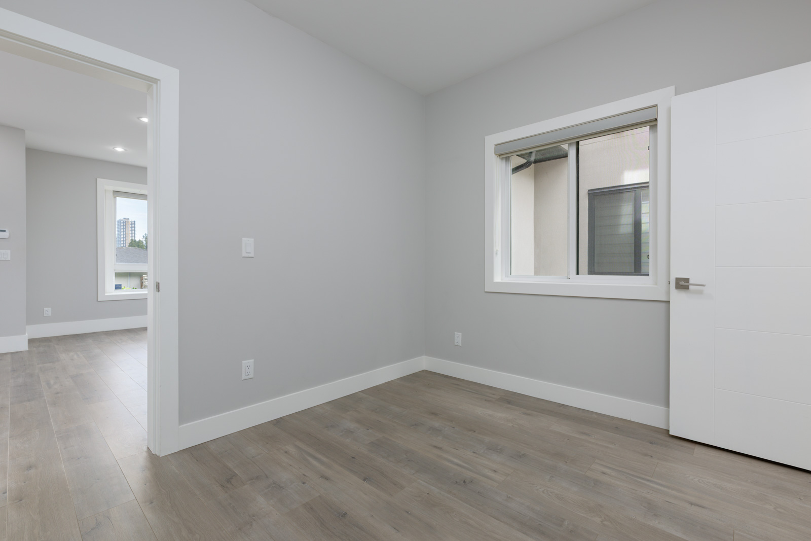 Empty room with light gray walls, a window, white trim, open white door, and wood floor; another room with windows is visible through the doorway.
