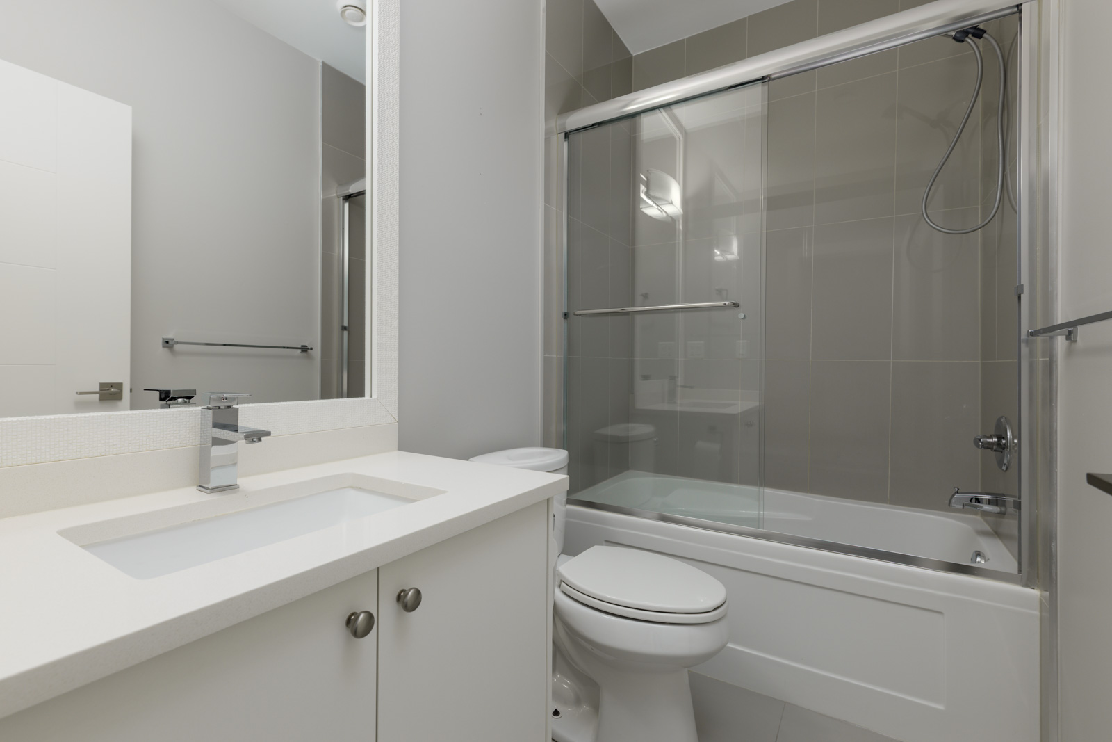 Modern bathroom with a white vanity, sink, toilet, and a bathtub with a glass sliding door and gray tiled walls.