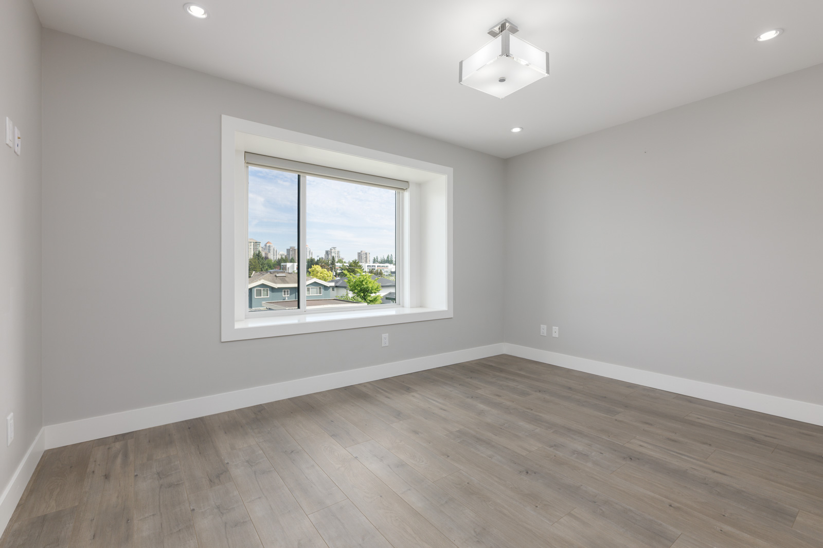Empty room with light gray walls, wood flooring, a large window with a view of houses and trees, and a modern ceiling light fixture.