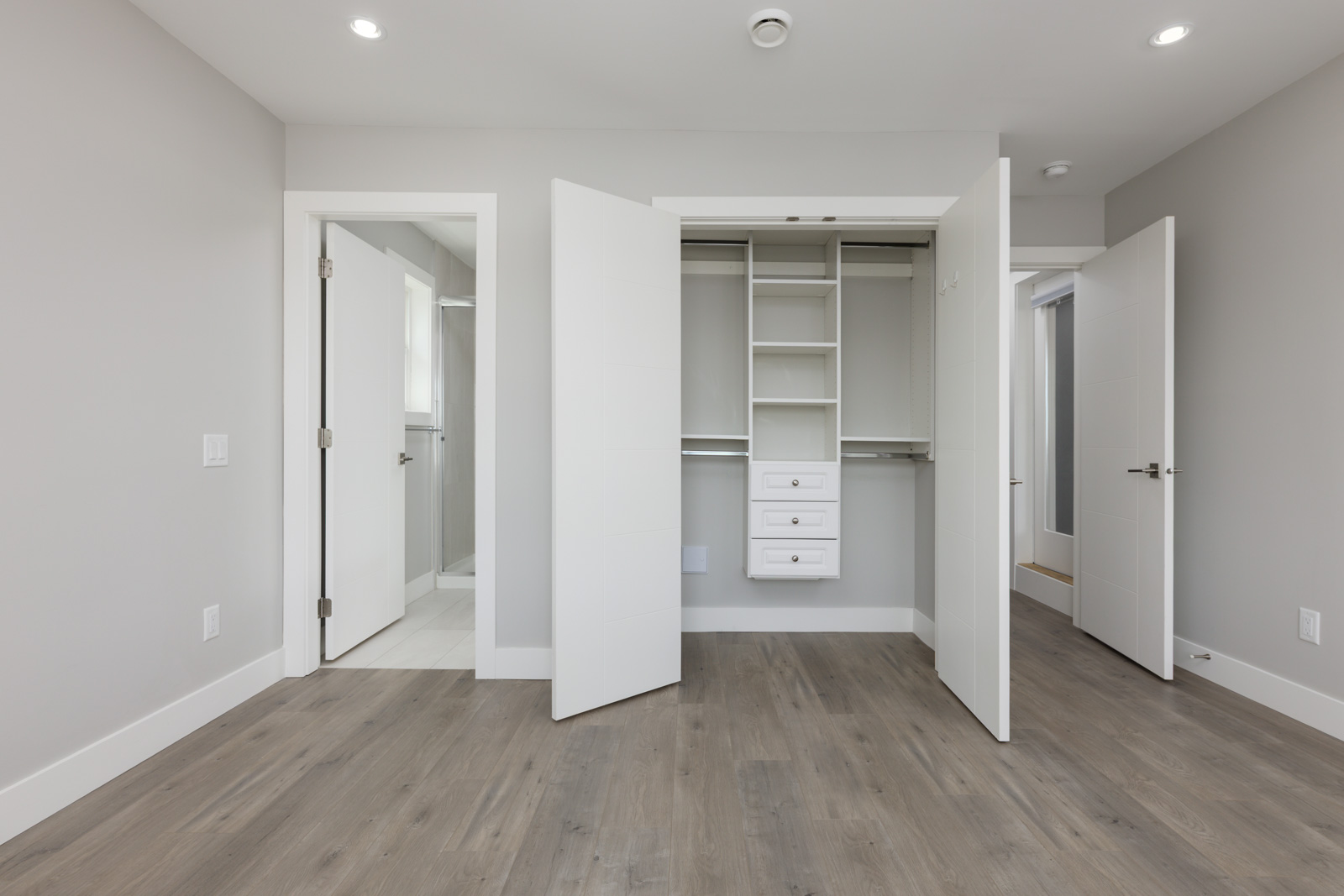 Empty room with light gray walls, wood flooring, an open closet with shelves and drawers, and two open white doors leading to other rooms.