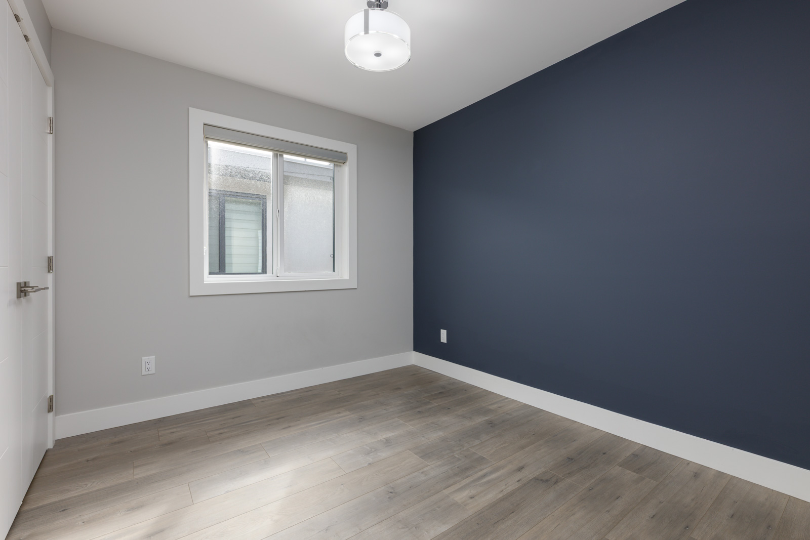 Empty room with light wood flooring, white trim, a large window, a white ceiling light, and two-tone walls in gray and dark blue.