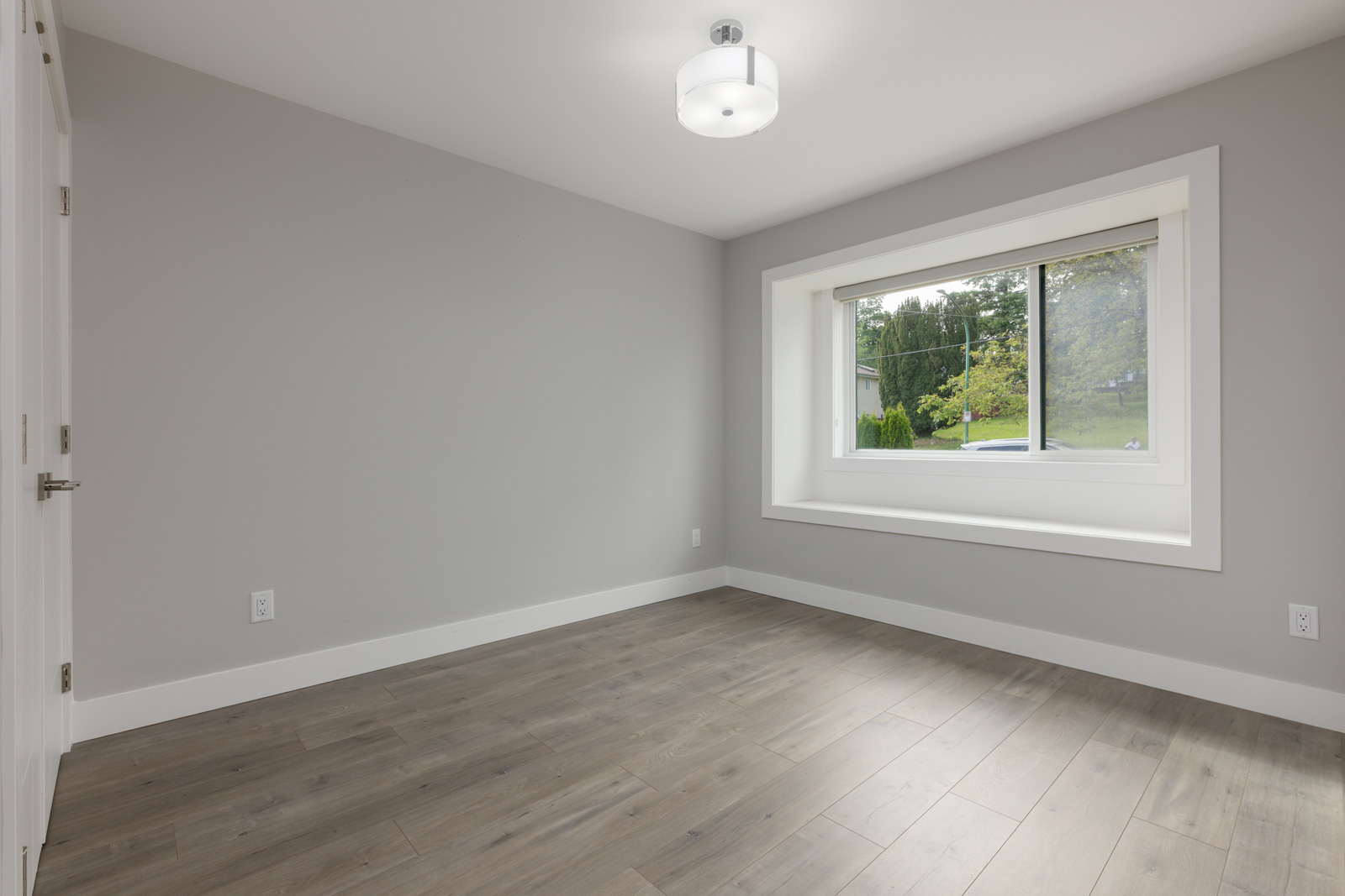 Empty room with light gray walls, wood flooring, a ceiling light fixture, and a large window with a white frame overlooking greenery.