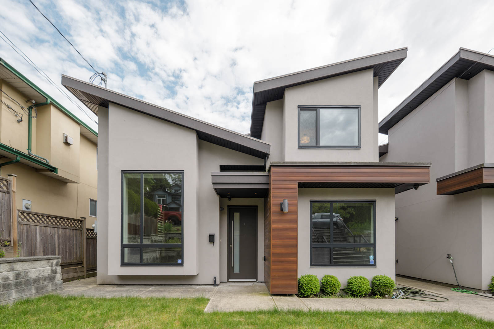 Modern two-story duplex with light gray exterior, large windows, wood accent panel, small front yard, and concrete walkway under a cloudy sky.