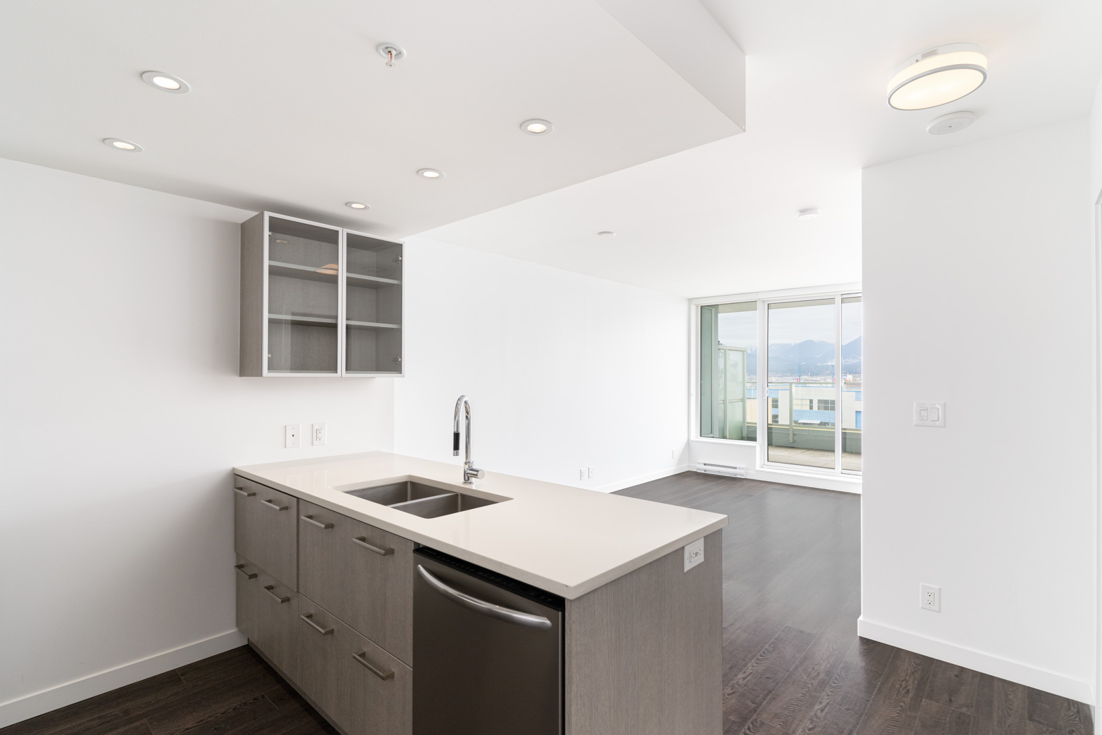 Modern kitchen with gray cabinets, white countertops, built-in sink, and dishwasher; open layout extends to a bright living area with large windows and mountain views.