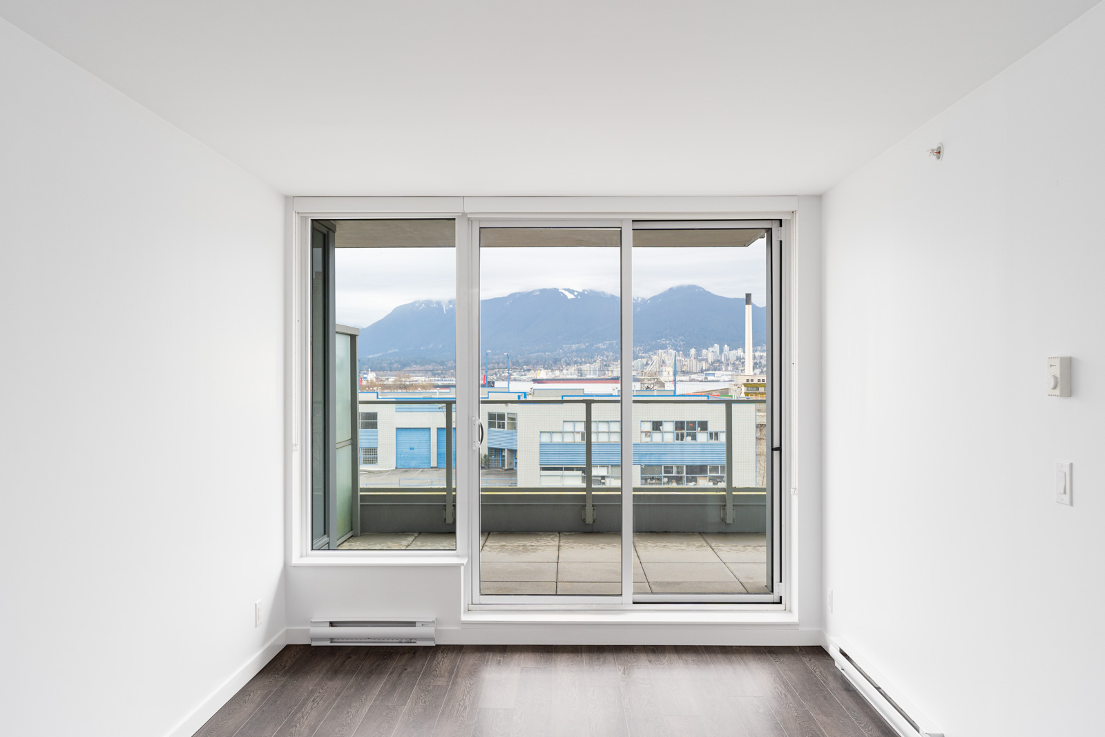 Empty white room with dark wood floor, large glass sliding doors leading to a balcony, and a view of city buildings and mountains in the background.