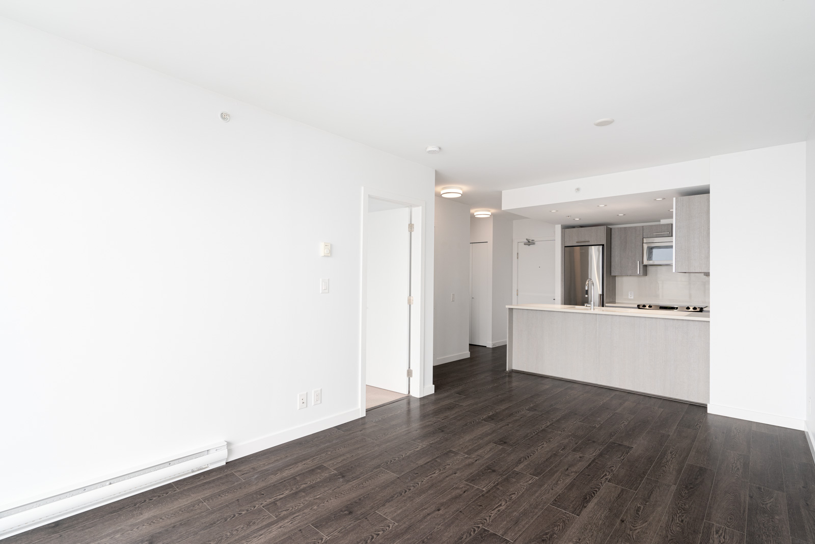 Unfurnished modern apartment with white walls, dark wood floors, and an open kitchen featuring light cabinets and stainless steel appliances.