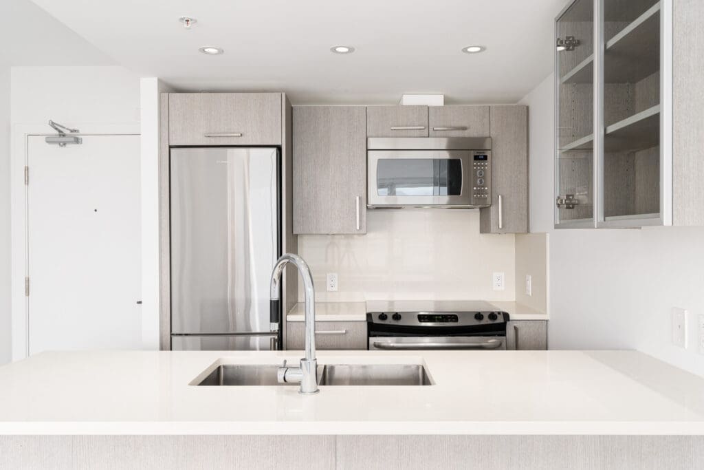 Modern kitchen with light wood cabinets, stainless steel appliances including a refrigerator, microwave, and stove, and a sink set in a white countertop island.
