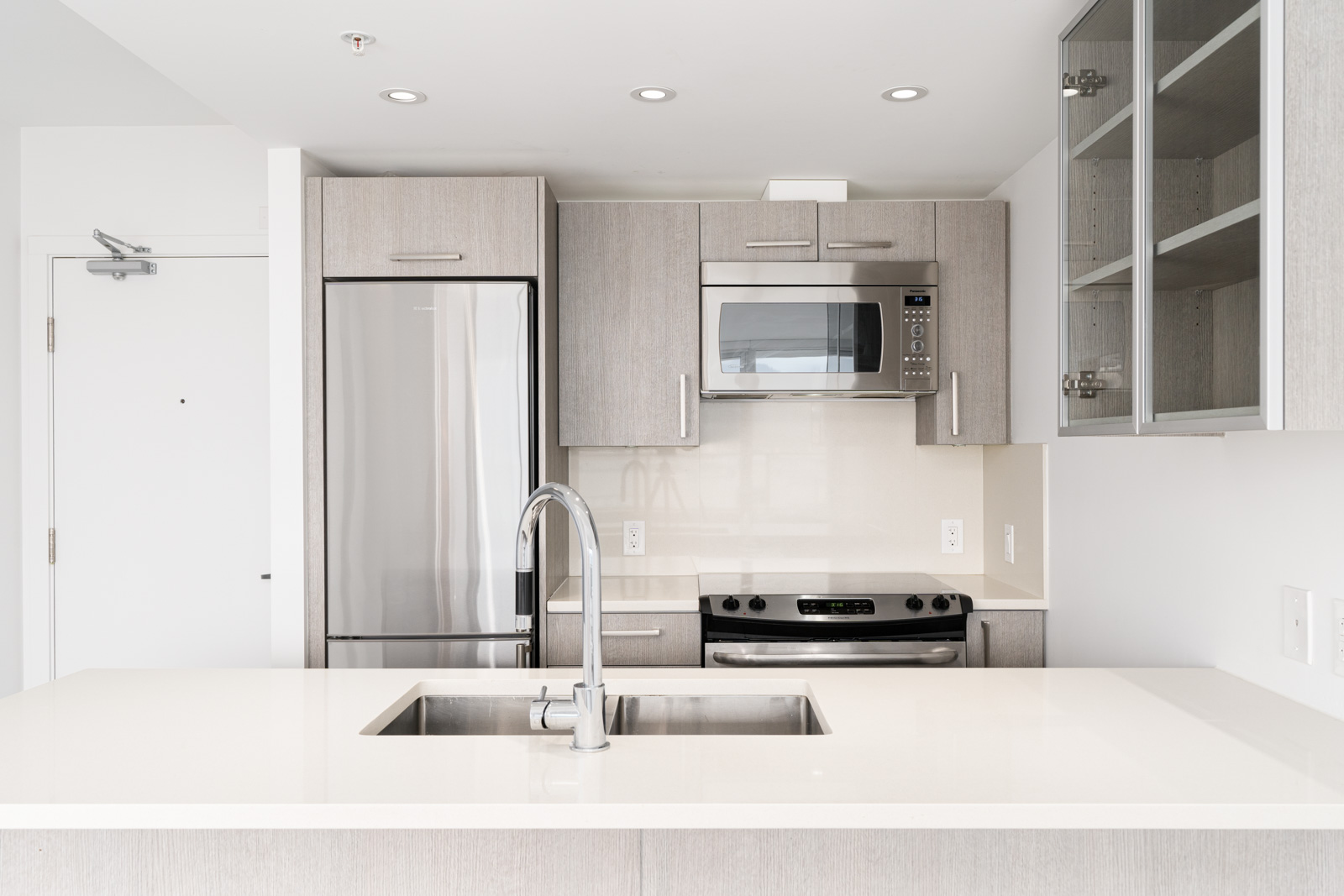 Modern kitchen with light wood cabinets, stainless steel appliances including a refrigerator, microwave, and stove, and a sink set in a white countertop island.