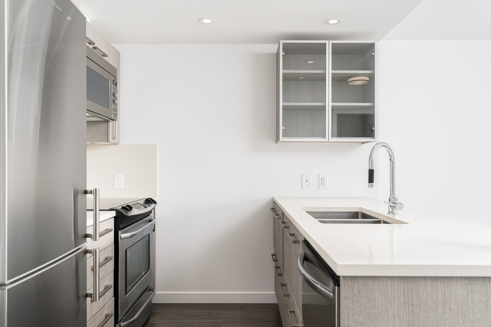 Modern kitchen with stainless steel appliances, light-colored cabinets, a built-in oven, glass-front upper cabinets, and a sink with a high-arc faucet on a white countertop.