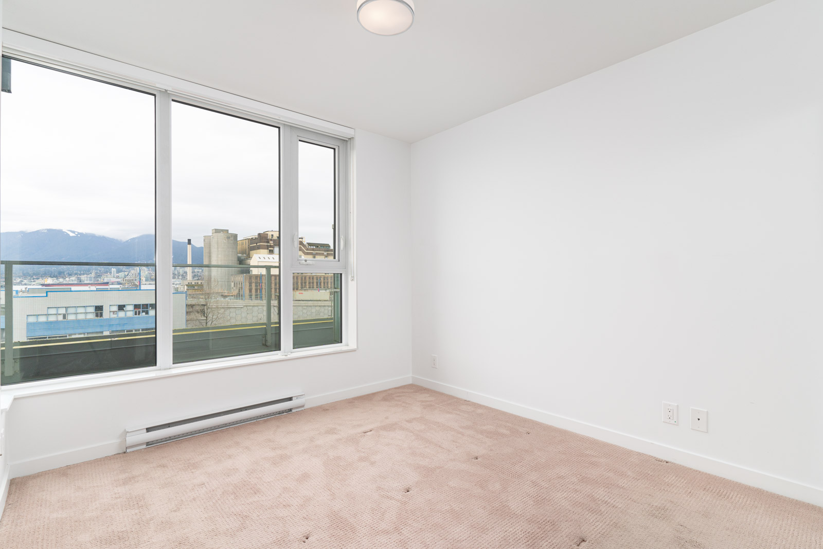 Empty room with beige carpet, white walls, and large windows offering a view of city buildings and mountains in the background.