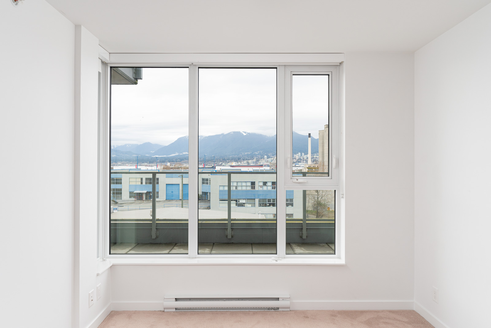 A large window in a white room overlooks buildings, a balcony, and distant mountains under an overcast sky.