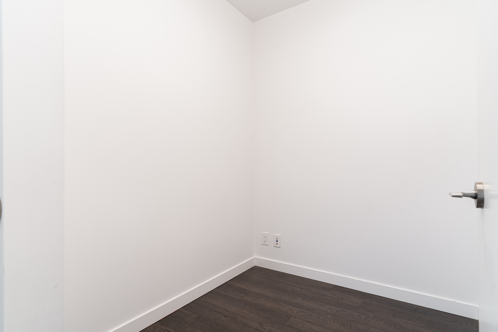 A small, empty room with white walls, dark wood flooring, a closed door, and two electrical outlets on the wall.