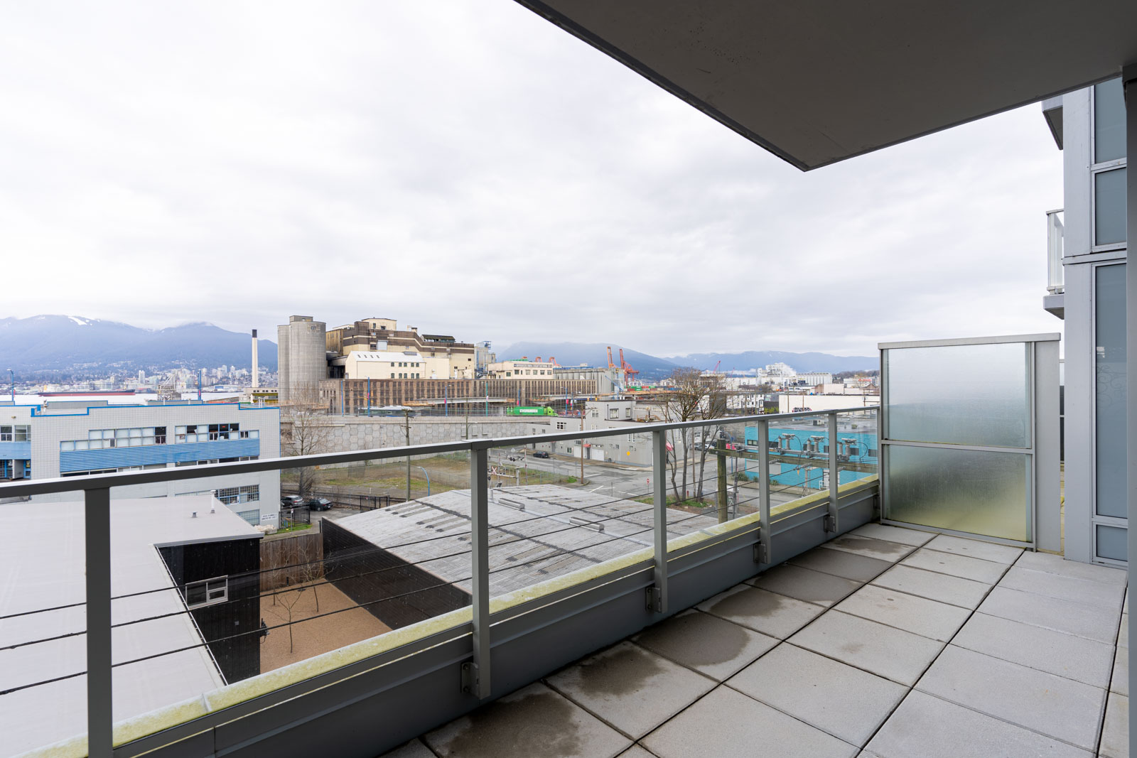 A spacious balcony with glass railings overlooks industrial buildings, mountains, and cloudy skies in the distance.