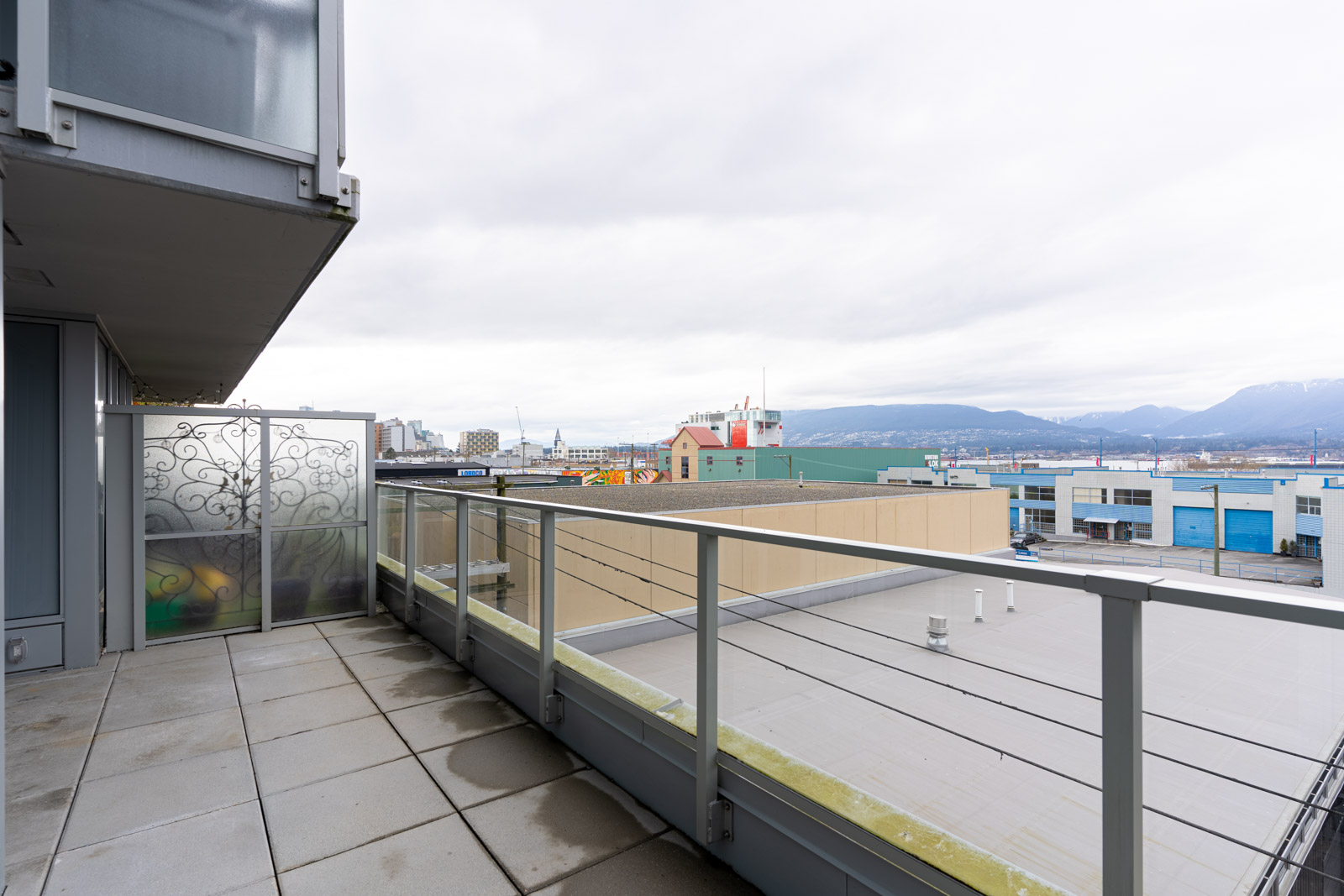 A view from a balcony with glass railing overlooking industrial buildings, with mountains visible in the background under a cloudy sky.