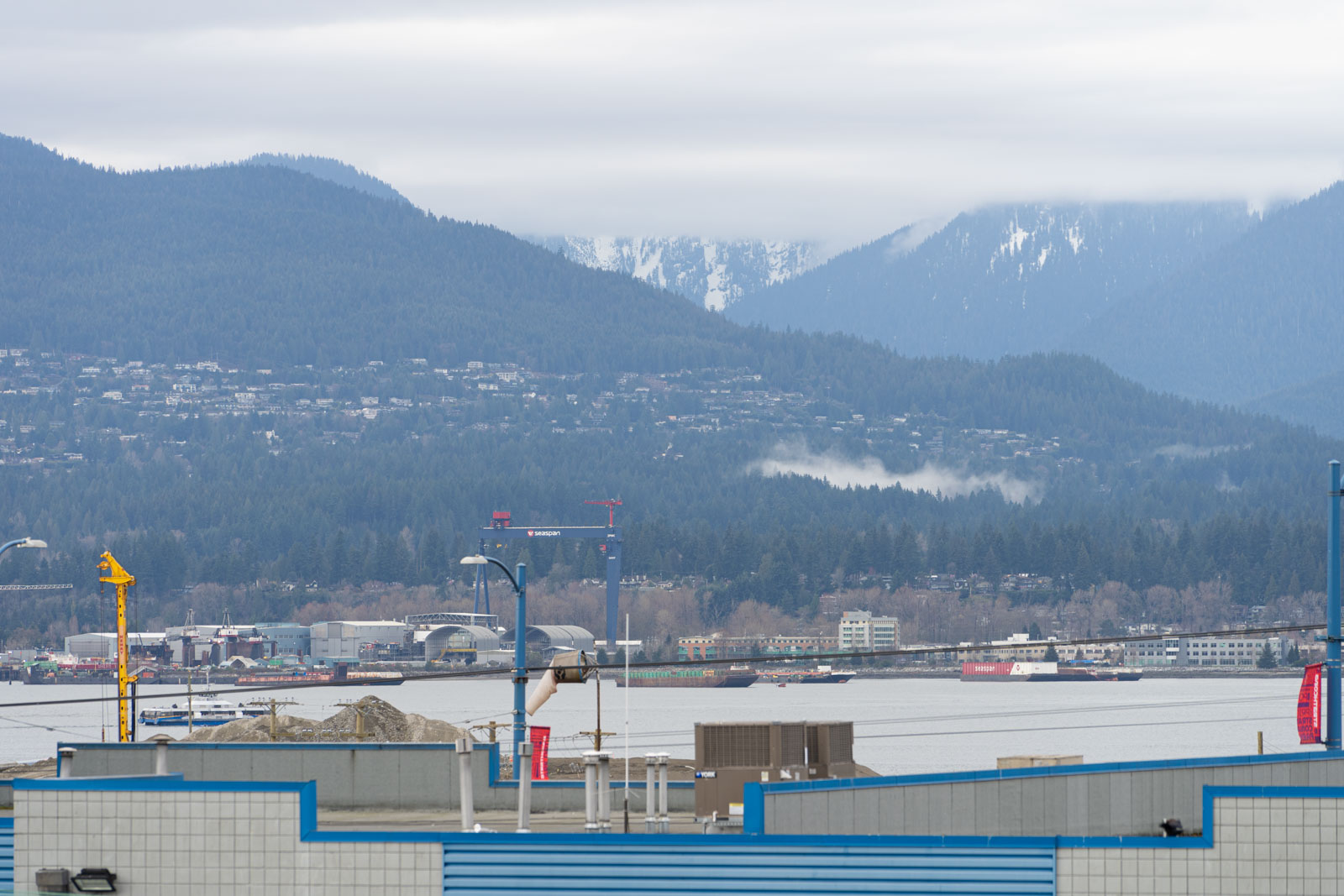 A view of a river with barges, a construction crane, industrial buildings in the foreground, and forested mountains in the background under a cloudy sky.