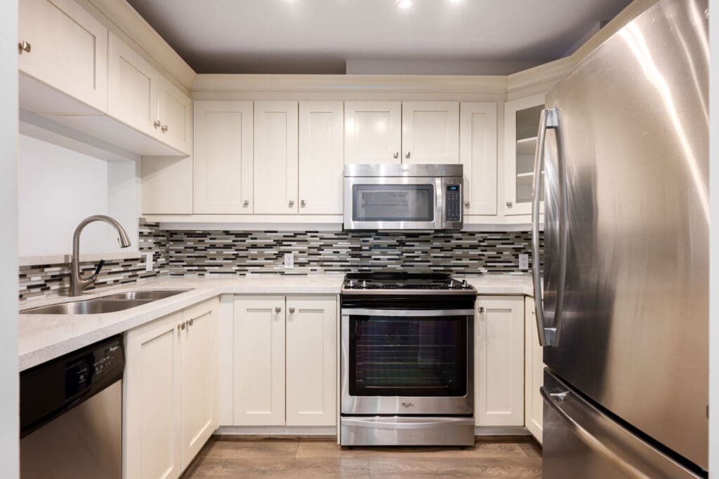 Modern kitchen with white cabinets, stainless steel appliances, a tile backsplash, and wood flooring. Features a double sink, stove, microwave, dishwasher, and refrigerator.