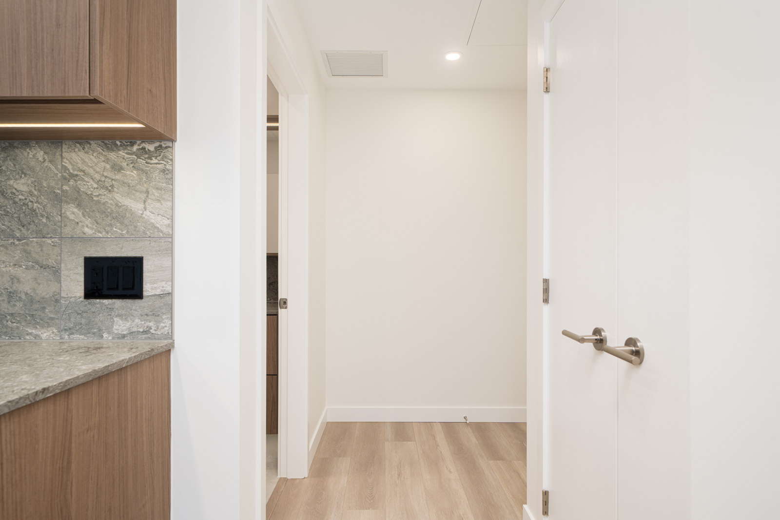 A hallway with light wood floors, white walls, and doors; a section of a kitchen counter with marble backsplash and cabinets is visible on the left.