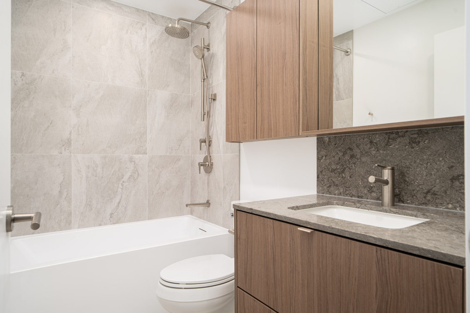 Modern bathroom with light stone tile walls, a bathtub with a rainfall showerhead, wood cabinetry, and a sink with a stone countertop and metal fixtures.