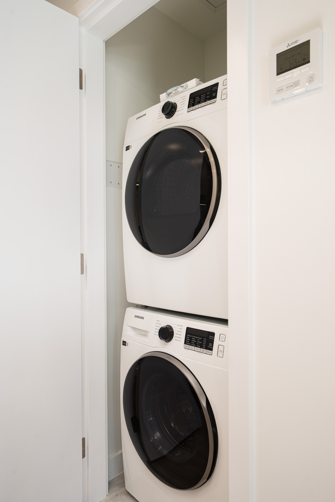 A stacked washer and dryer are positioned in a small laundry closet with a white door, next to a digital thermostat on the wall.