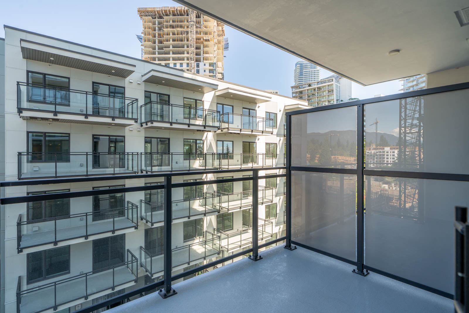 A view from a modern apartment balcony with glass panels, overlooking a neighboring apartment building and a construction site in the background.