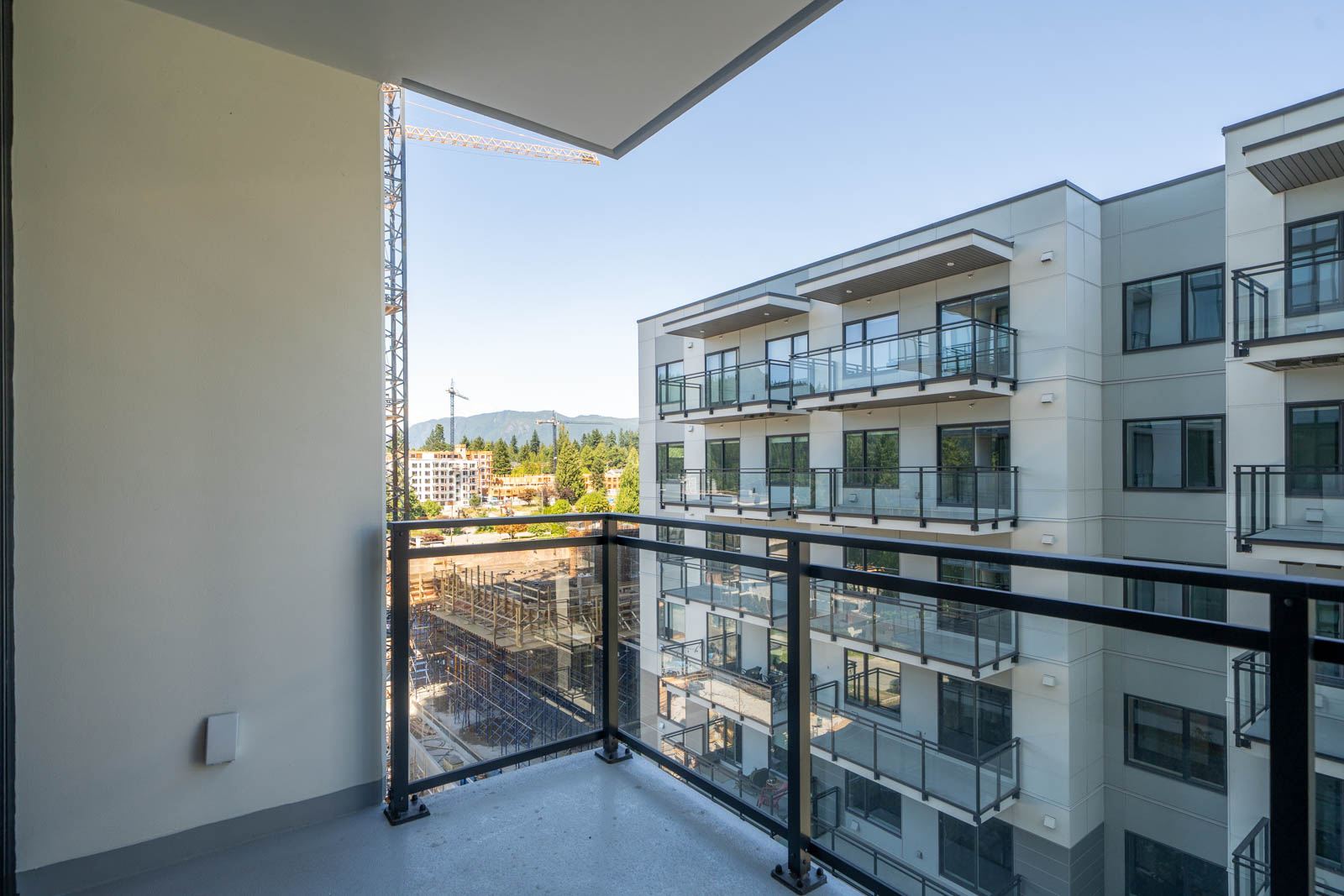 A modern apartment balcony with glass railing overlooks a construction site, an adjacent building, trees, and distant mountains under a clear sky.