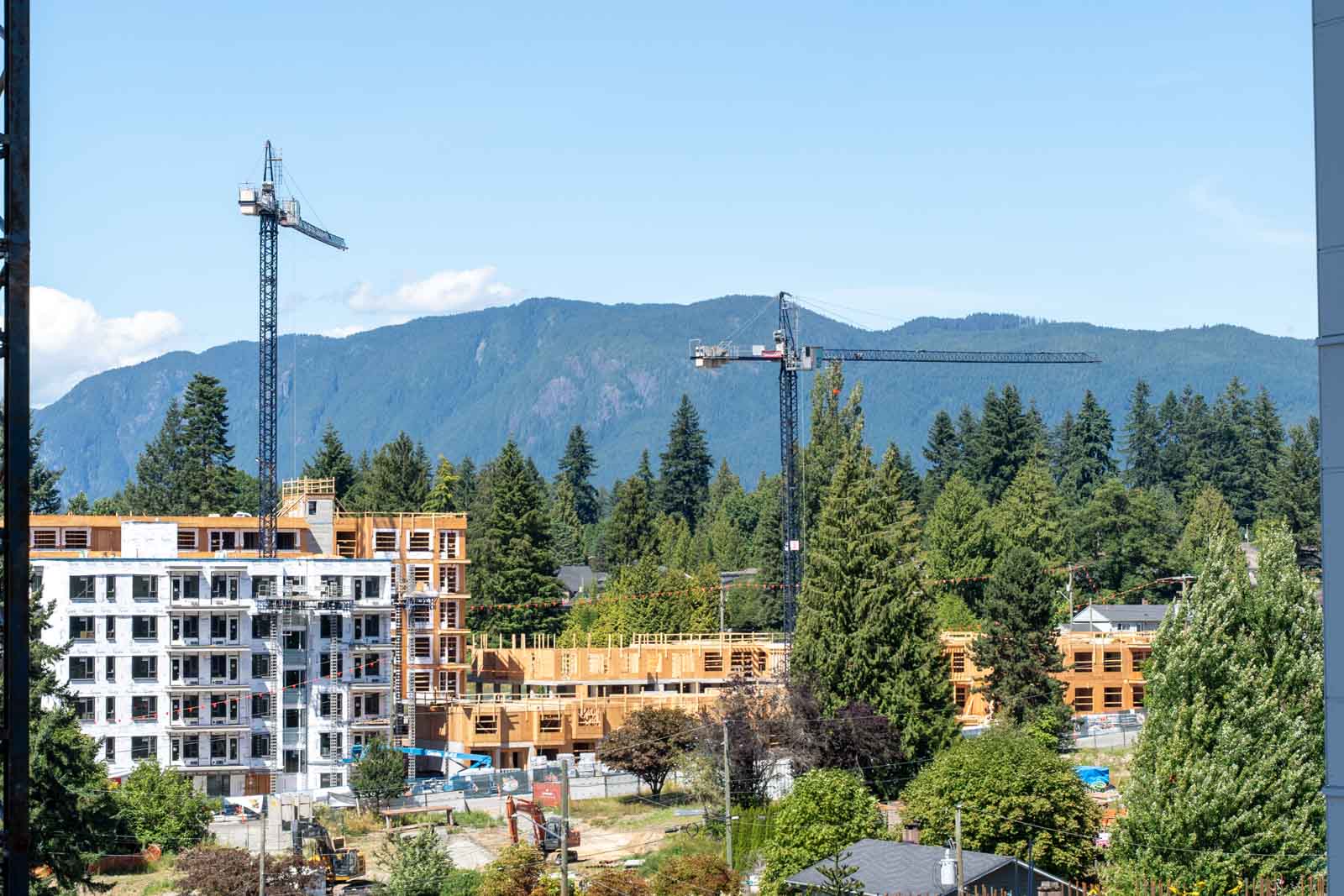 Construction site with two tower cranes and partially built multi-story buildings, surrounded by trees and mountains in the background under a clear sky.