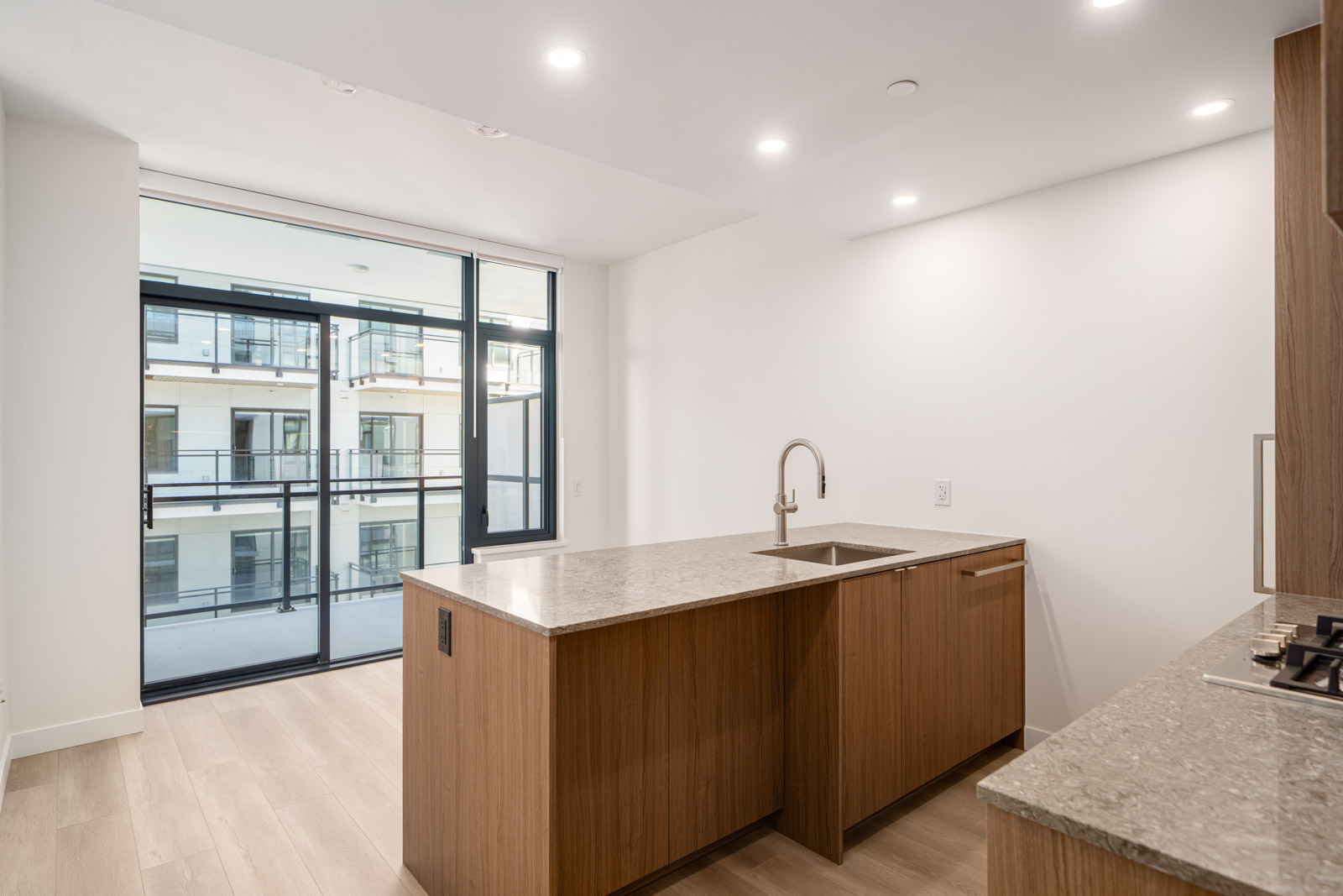 Modern kitchen with wooden cabinets, stone countertops, a sink island, and large glass doors leading to a balcony with a view of adjacent apartment buildings.